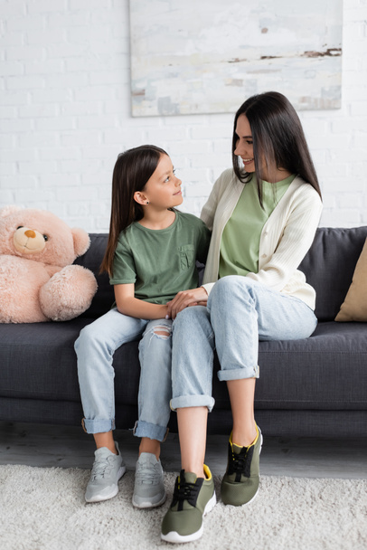 full length of girl and nanny sitting on couch near teddy bear and smiling at each other - Zdjęcie, obraz