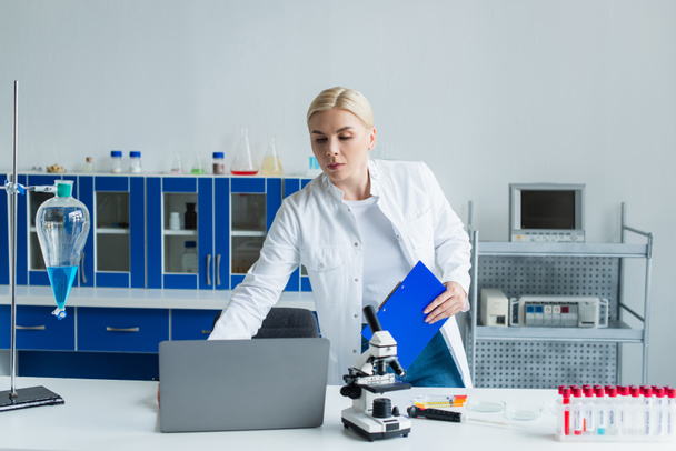 Scientist holding clipboard near laptop and test tubes in lab  - Foto, Imagen