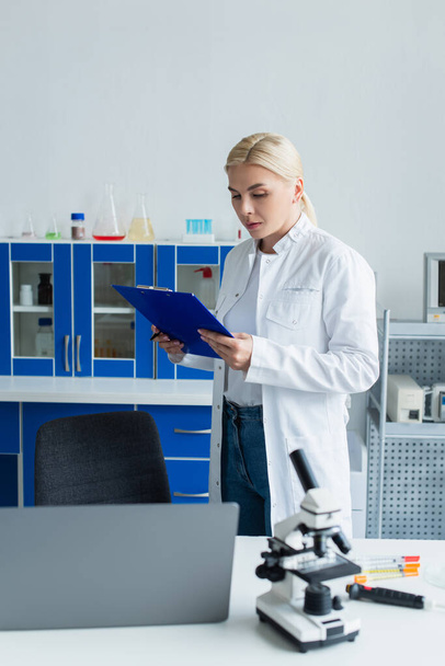 Scientist holding clipboard near laptop and microscope in lab  - Фото, изображение