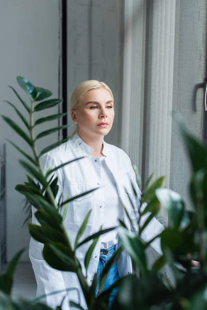 Блондинка в белом халате стоит рядом с растением в лаборатории - Фото, изображение
