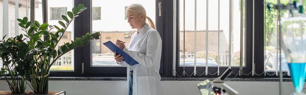 Side view of scientist writing on clipboard near plants and blurred flask in lab, banner  - Фото, зображення