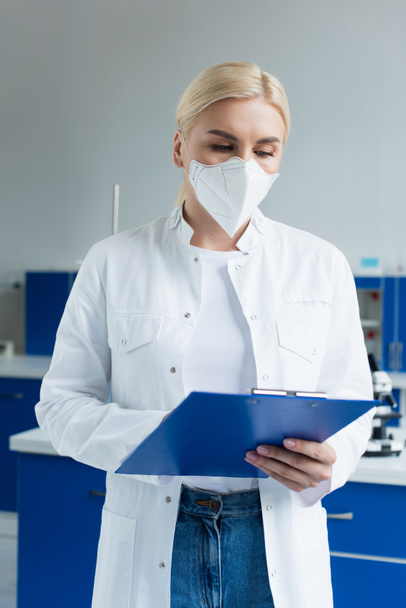 Scientist in white coat and protective mask holding clipboard while working in lab - Photo, Image