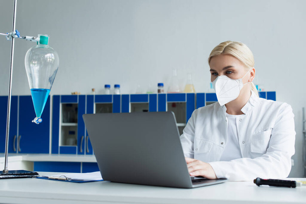 Вчений в захисній масці використовує ноутбук біля колби в лабораторії
  - Фото, зображення