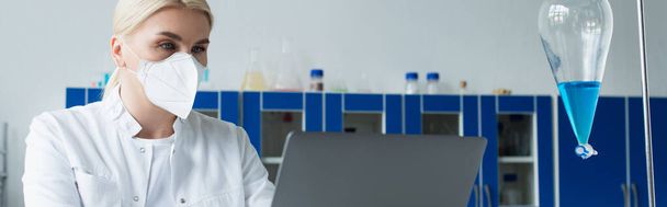 Cientista loira em máscara protetora e casaco branco usando laptop perto de frasco com líquido em laboratório, banner  - Foto, Imagem
