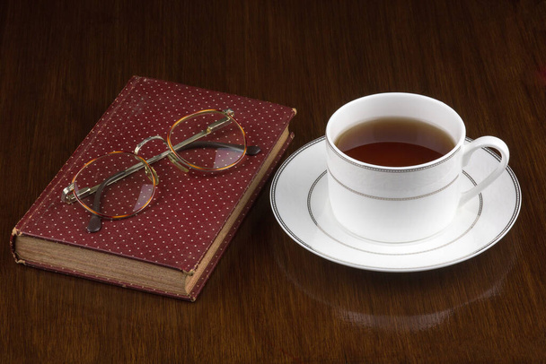 Чашка чаю зі старою книгою та окулярами на старій полірованій дерев'яній поверхні
 - Фото, зображення