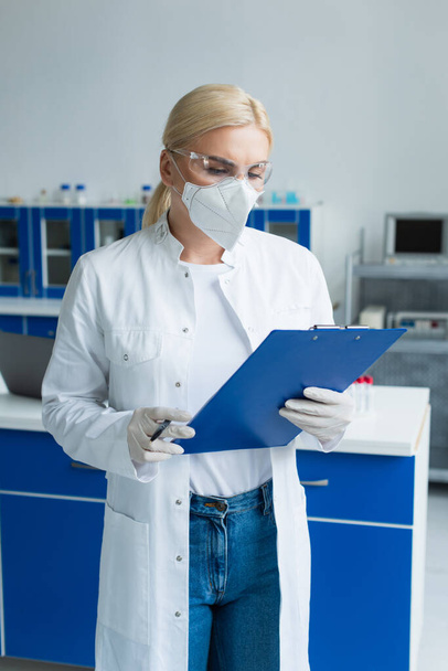 Blonde scientist in protective mask holding clipboard in laboratory  - Foto, Imagen