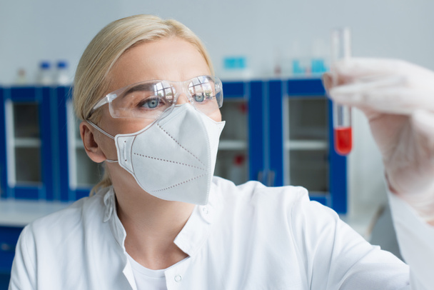 Blonde scientist in goggles and protective mask holding blurred test tube in lab  - Foto, Imagen