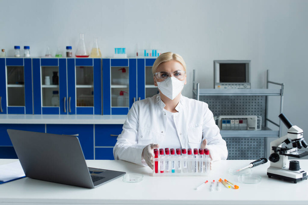 Scientist in protective mask looking at camera near test tubes and microscope in lab  - Foto, Imagen