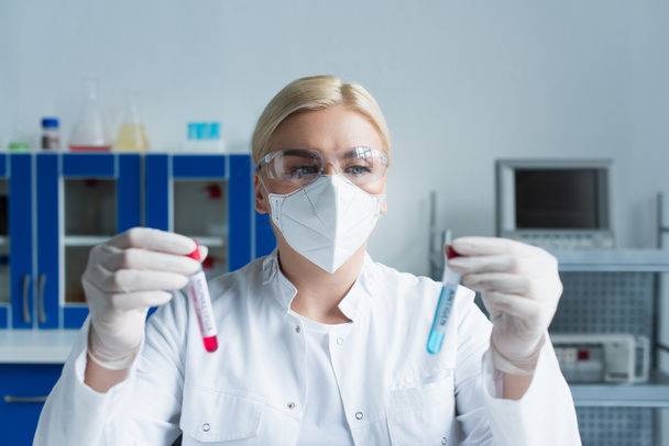 Cientista em máscara protetora segurando tubos de ensaio desfocados em laboratório  - Foto, Imagem