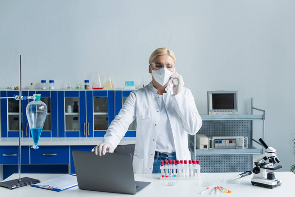 Scienziato in maschera e occhiali che parla su smartphone vicino a provette e microscopio in laboratorio  - Foto, immagini