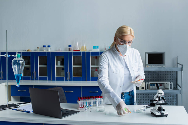 Scientist in protective mask holding syringes near test tubes and laptop in lab  - Foto, Bild