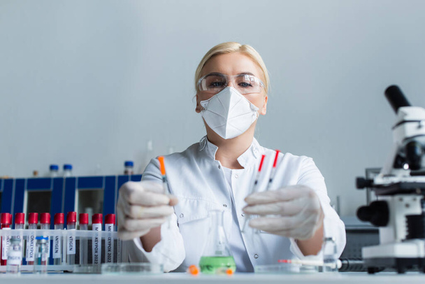 Low angle view of scientist in mask holding blurred syringes near microscope and test tubes in lab  - Photo, image