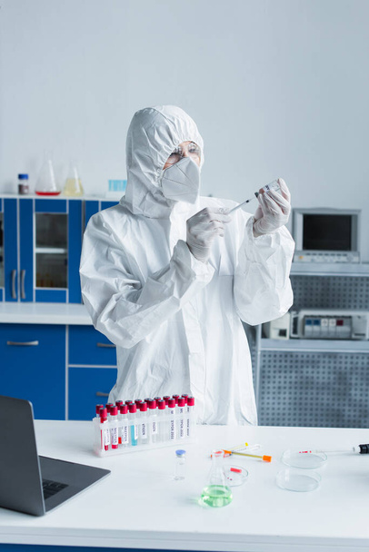 Naukowiec w kombinezonie ochronnym trzymający strzykawkę i szczepionkę w pobliżu probówek w laboratorium  - Zdjęcie, obraz