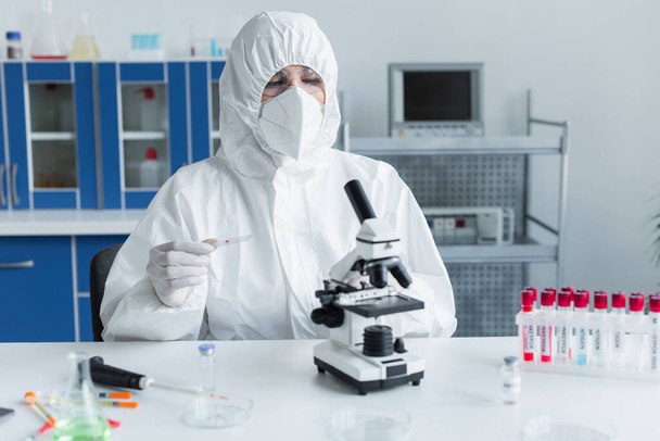 Scientist in hazmat suit holding glass near microscope and test tubes in lab  - Zdjęcie, obraz