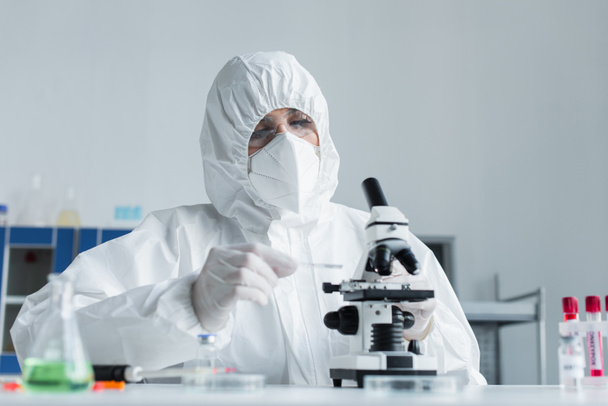 Wissenschaftler im Schutzanzug hält Glas, während er im Labor mit dem Mikroskop arbeitet - Foto, Bild