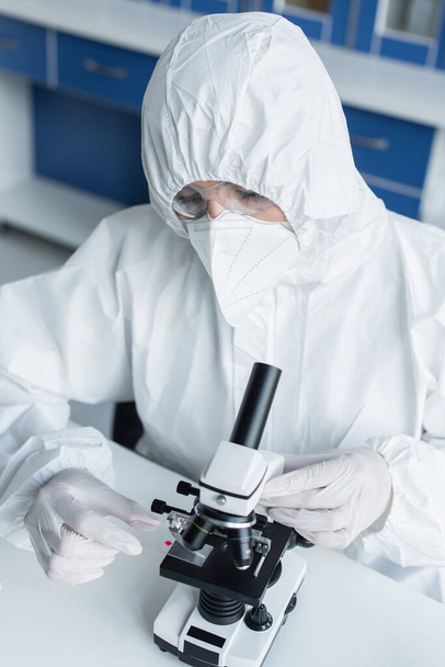 Scientist in hazmat suit holding glass near microscope in lab  - Fotó, kép
