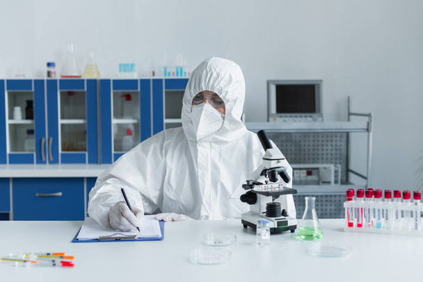 Scientist in hazmat suit writing on clipboard near microscope and test tubes in lab  - Foto, afbeelding