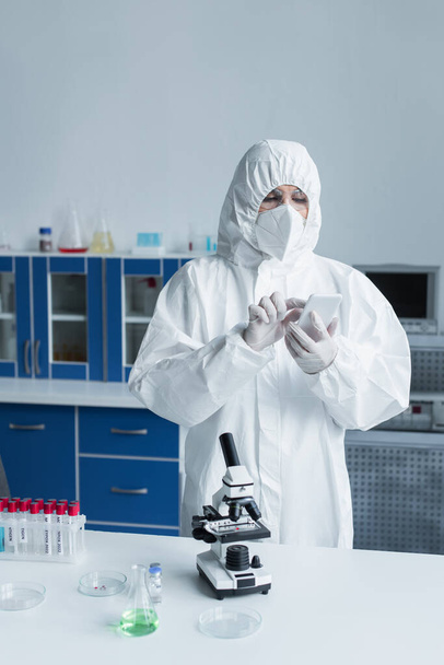 Scientist in hazmat suit using smartphone near microscope and test tubes in lab  - Foto, Imagen