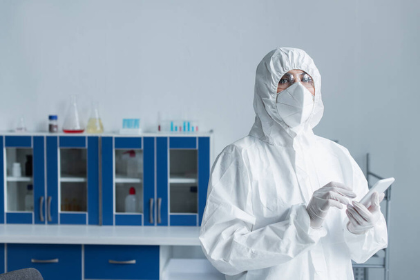 Wetenschapper in beschermende pak en bril met smartphone en kijken naar de camera in het laboratorium - Foto, afbeelding