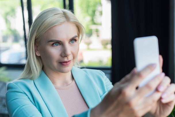 Blonde psychologist having video call on blurred smartphone in consultation room  - Foto, Bild