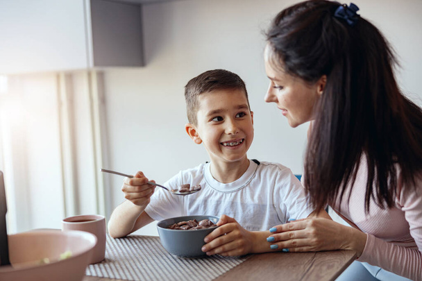 Portrait, small teen boy eating cereals with milk in kichen and looking at mother. Having breakfast. - Foto, Imagem