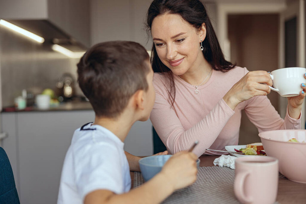 Mãe muito feliz olhando para o pequeno filho adolescente e sorrindo, bebendo café de manhã. Café da manhã. - Foto, Imagem