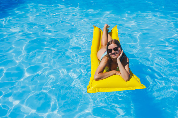 Hübsche junge Frau in Badeanzug und Sonnenbrille, die auf gelben aufblasbaren Matratzen im Pool liegt. Genießen Sie Sonnenbaden und Entspannung. - Foto, Bild