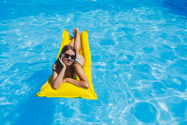 Beautiful young woman in sunglasses on a yellow inflatable mattress in a swimsuit swims in the pool of a luxury hotel, summer vacation, travel, smile - Foto, Imagem