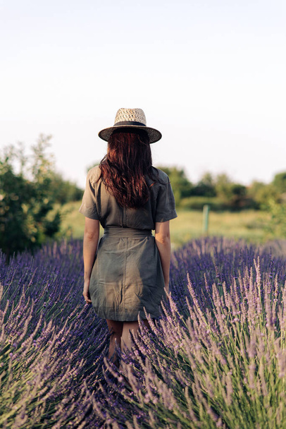 Young woman in hat walks among lavender field and enjoys by blooming. Back view against sky. - Fotoğraf, Görsel
