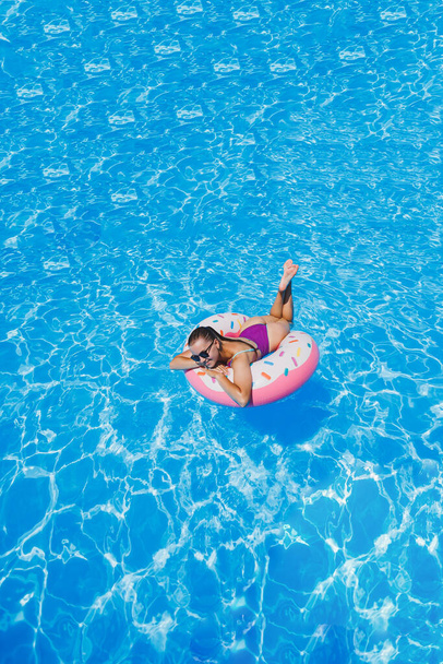 Beautiful slim young woman in a swimsuit enjoying the water park floating in an inflatable big ring on a sparkling blue pool smiling at the camera. Summer vacation. - Foto, Imagem