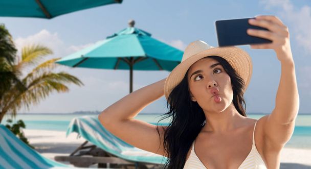 travel, tourism and summer vacation concept - happy smiling young woman in bikini swimsuit and straw hat taking selfie with smartphone over tropical beach background in french polynesia - 写真・画像
