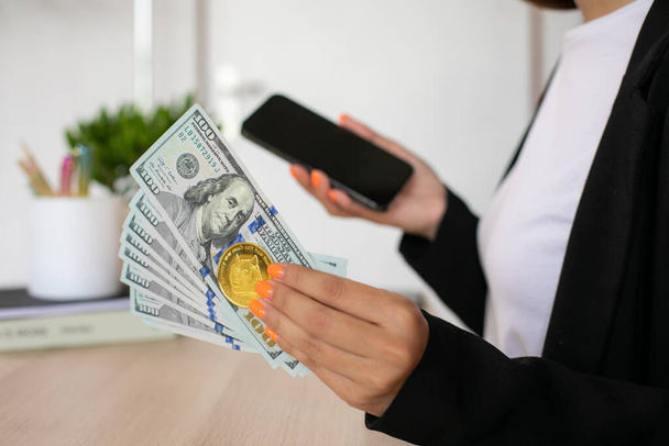 Billetes de dólar y dogecoin a manos de la mujer con teléfono en el lugar de trabajo. Foto de alta calidad - Foto, Imagen