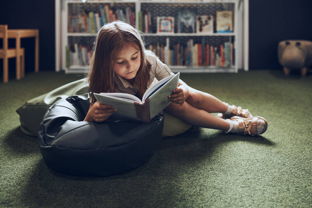 Schoolgirl reading book in school library. Primary school pupil is involved in book. Child doing homework. Smart girl learning from book. Benefits of everyday reading. Child curiosity - Foto, Imagen