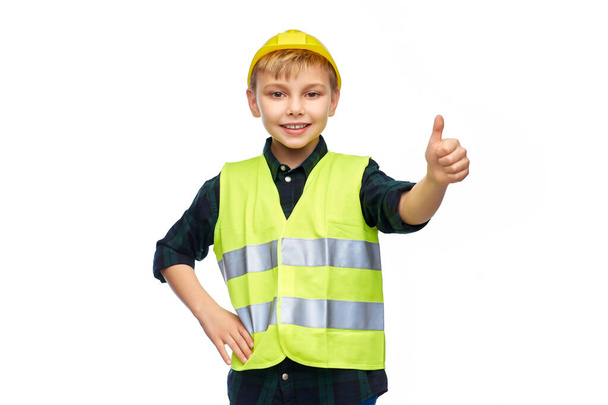 κτίριο, κατασκευή και επάγγελμα έννοια - χαρούμενος χαμογελαστό μικρό αγόρι σε προστατευτικό κράνος και γιλέκο ασφαλείας δείχνει αντίχειρες πάνω από λευκό φόντο - Φωτογραφία, εικόνα