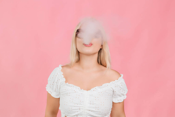 молодая светловолосая девушка в очках выдыхает дым на розовом фоне - Фото, изображение