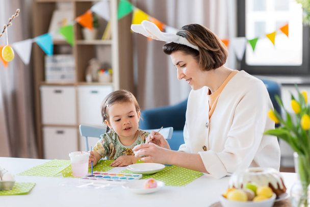 holidays, family and people concept - happy mother and little baby daughter coloring easter eggs with watercolors at home - Φωτογραφία, εικόνα