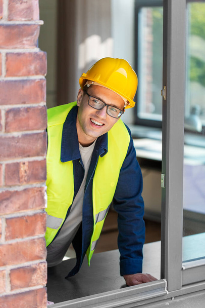 construction business and building concept - smiling male builder in helmet and safety west looking out window - Photo, image