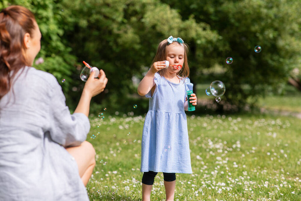 family, motherhood and people concept - happy mother with little daughter blowing soap bubbles at summer park or garden - Foto, Imagen