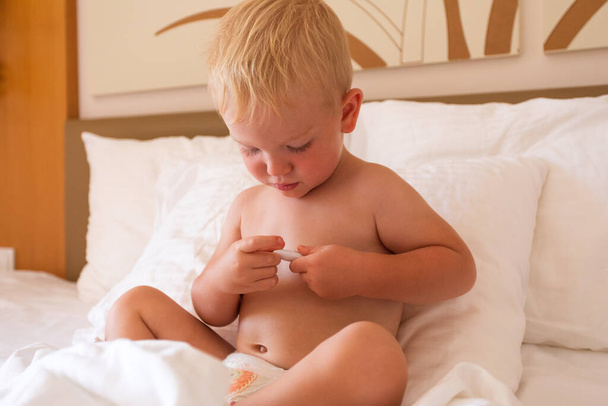 Egy kisfiú leül az ágyba, egy hőmérőt tart a kezében, és megnézi a lázát. - Fotó, kép