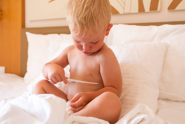 A little boy sits in bed and holds a thermometer in his hands and looks at his temperature - Foto, Bild