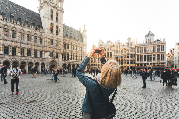A young woman stands with her back to the camera and takes pictures on her phone for social networks against the background of the Grand Place in Brussels, Belgium - Foto, Imagem