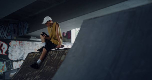 Стильний чоловік переглядає Інтернет на мобільному телефоні в скейтпарку. Молодий ковзаняр прокручує серфінг у соціальних мережах смартфон, що сидить на рампі. Технологія цифрового гаджета концепція міської субкультури
. - Фото, зображення
