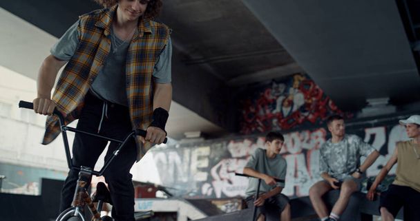 Active man practicing on bmx bicycle at skate park with graffiti wall background. Bmx rider training trick outdoors. Teenage friends meeting at skatepark. Healthy lifestyle urban subculture concept.  - Photo, Image