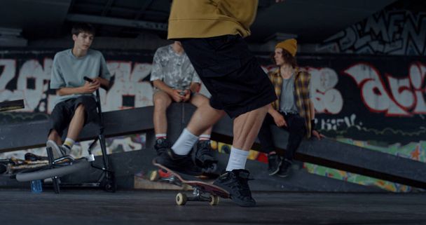 Millennial Boy bei Skateboard-Tricks im Freien. Aus nächster Nähe springen unbekannte Männerbeine auf ein Skateboard. In Großaufnahme betrachten jugendliche Hipster einen Typen mit Skateboard, der auf einem Skatepark sitzt. Lifestyle-Konzept - Foto, Bild