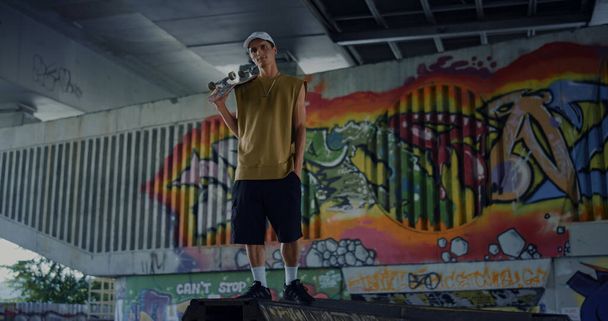 Χαλαρός άνθρωπος που κάνει διάλειμμα μετά το σκέιτμπορντ στο skatepark. Όμορφος σκέιτερ ποζάρει κάμερα έξω. Έφηβος που κρατάει σκέιτμπορντ στους ώμους του σε φόντο γκράφιτι. Η έννοια του τρόπου ζωής της νεολαίας.  - Φωτογραφία, εικόνα