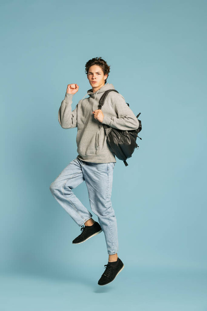 Ready to studying. Happy student in jeans and grey hoodie jumping isolated over blue background. Concept of emotions, facial expression, youth. Back to school - Fotó, kép