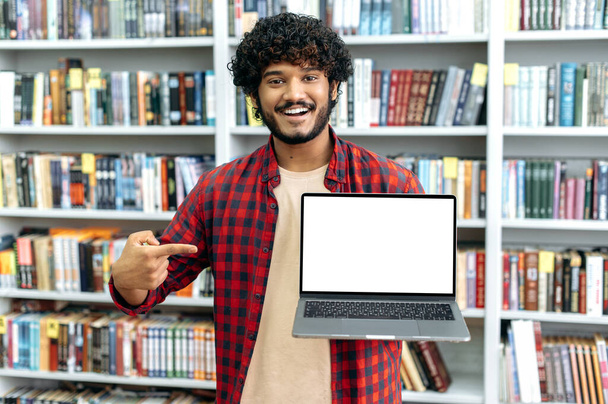 Positive friendly millennial male student, shows a laptop with a blank white mock-up screen, points fingers at it, stands in the library against the background of bookshelves, looks at camera, smiles - Foto, Imagen