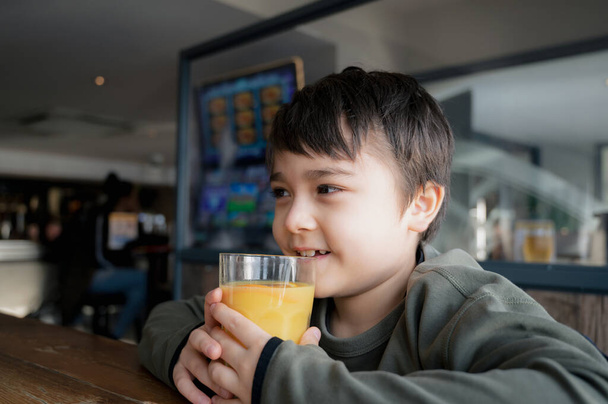 Side view Portrait Young kid drinking fresh orange juice for breakfast in cafe, Happy child boy drinking glass of fruit juice while waiting for food in restaurant, Healthy food lifestyle concept - Fotografie, Obrázek