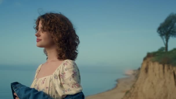Dreamy cute girl standing alone on empty sand beach quiet evening close up. Portrait of beautiful curly woman looking camera with calm smile. Attractive young lady relax on nature enjoying seascape. - Filmmaterial, Video