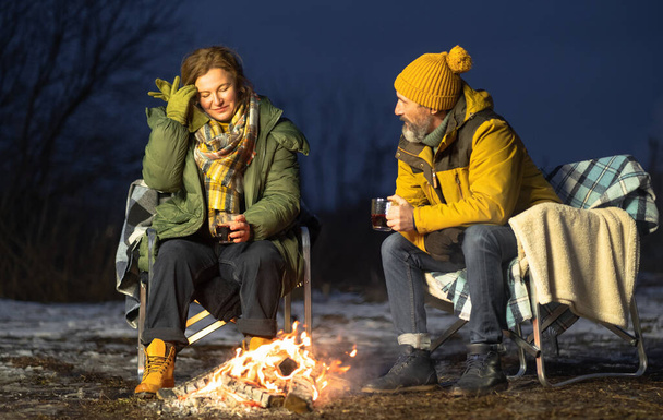 Middle aged couple sitting at campfire in snow. In love mature couple warming up at campfire drinking hot tea sitting on the plaid blankets in winter journey. Family vacation and journey concept.  - Φωτογραφία, εικόνα